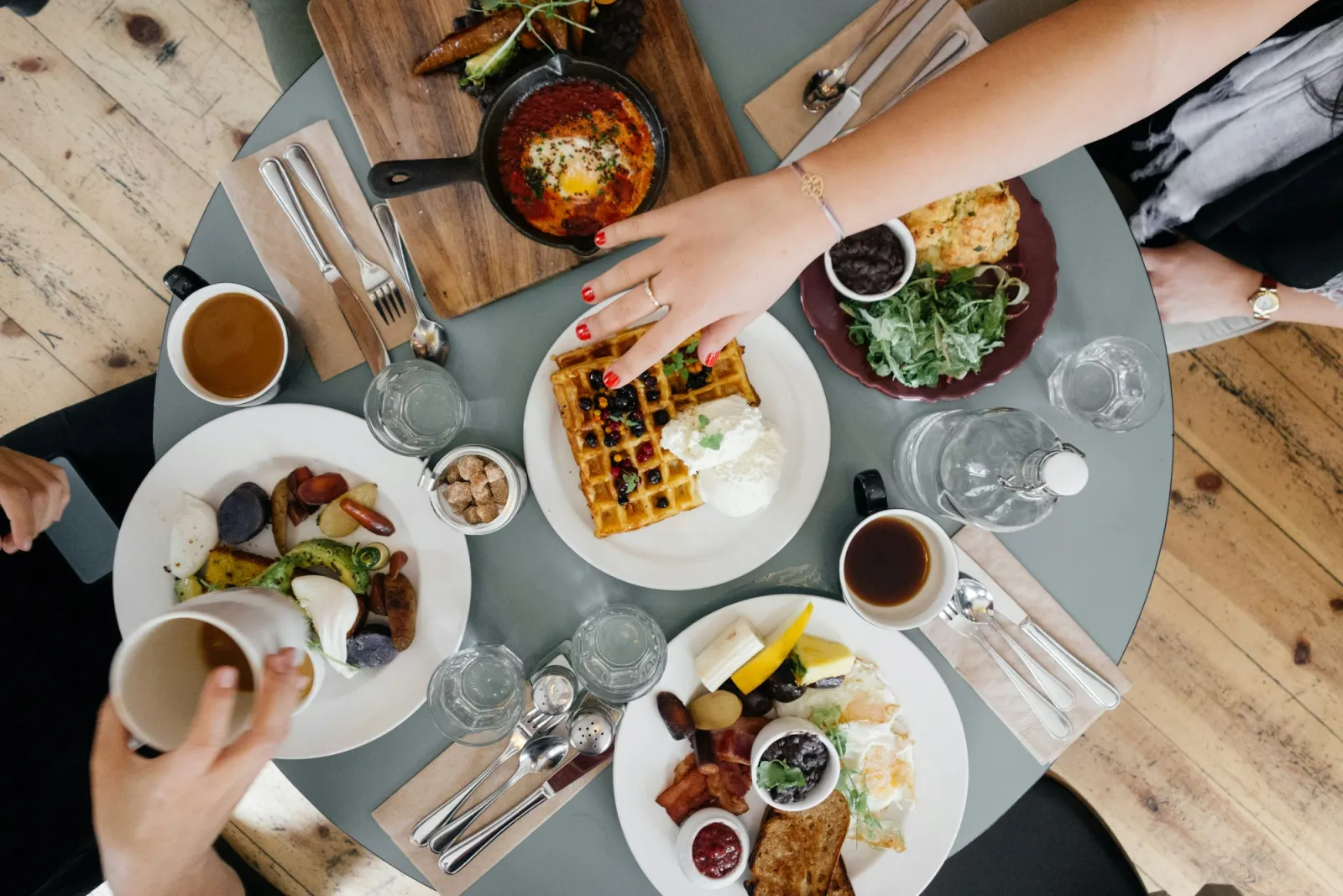 Friends sharing a warm brunch table from above, hands reaching for waffles and coffee at a restaurant